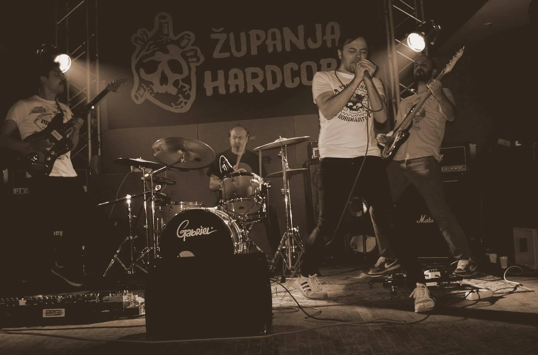 A four-member band performing on stage with a backdrop that says 'Županjska Hardcorg.' The band has a vocalist, two guitarists, and a drummer, with musical equipment including guitars, drums, and amplifiers.