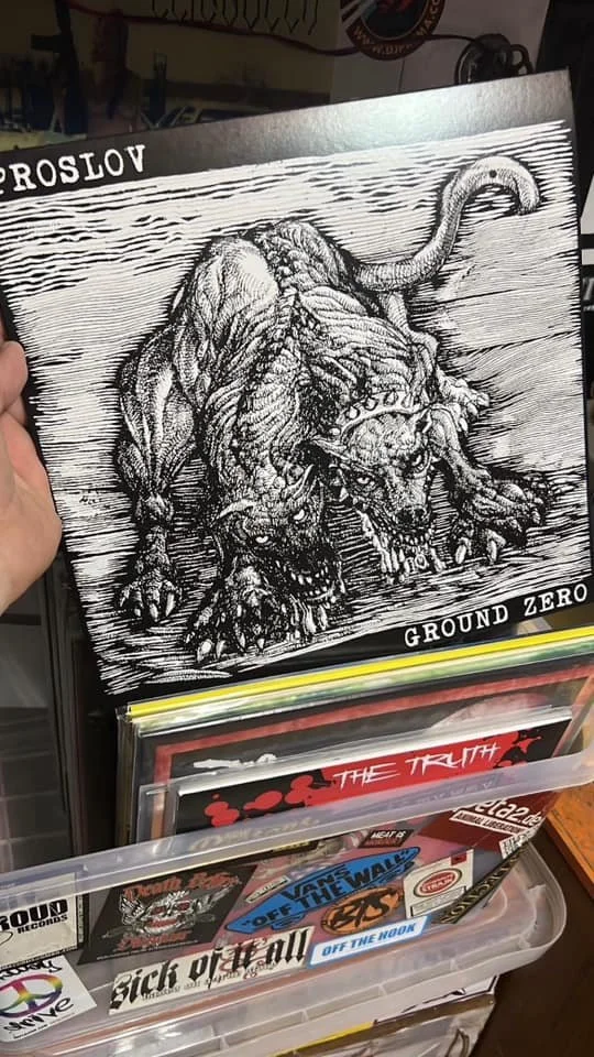 A black and white illustration of a fierce, snarling, large dog with a spiked collar, shown on a display rack among other records and artwork.