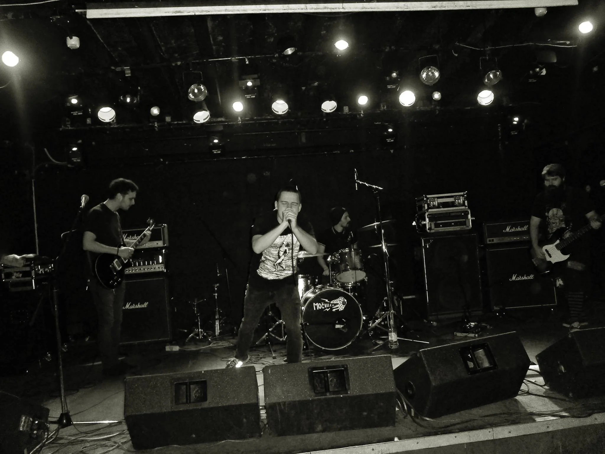 A band performing on stage with several musicians playing guitars and drums, and a lead singer holding a microphone. The stage has stacked amplifiers and stage lighting.