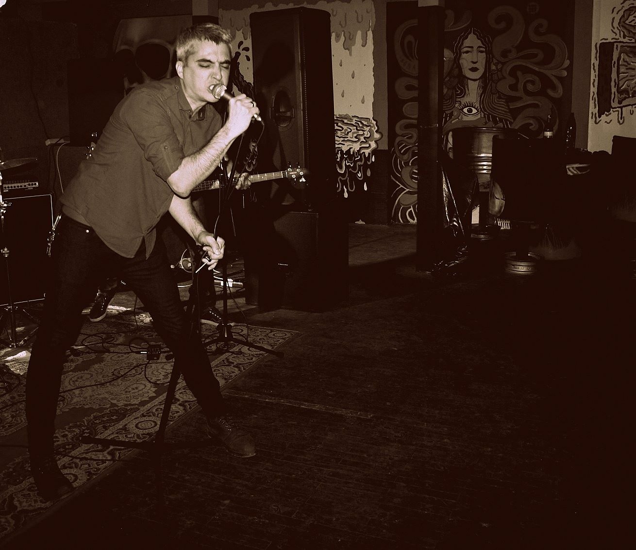 A young man passionately singing or speaking into a microphone on a stage with musical instruments and artwork in the background, in a dimly lit performance venue.