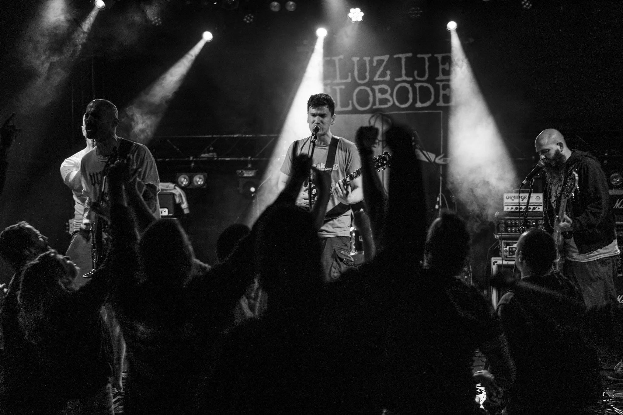 Black and white photo of a live music performance with three band members on stage. The central figure appears to be singing or speaking into a microphone while playing guitar. The stage is lit with spotlights and there is a crowd of people in the foreground. A sign with the words 'Luzije Slobode' is visible in the background.