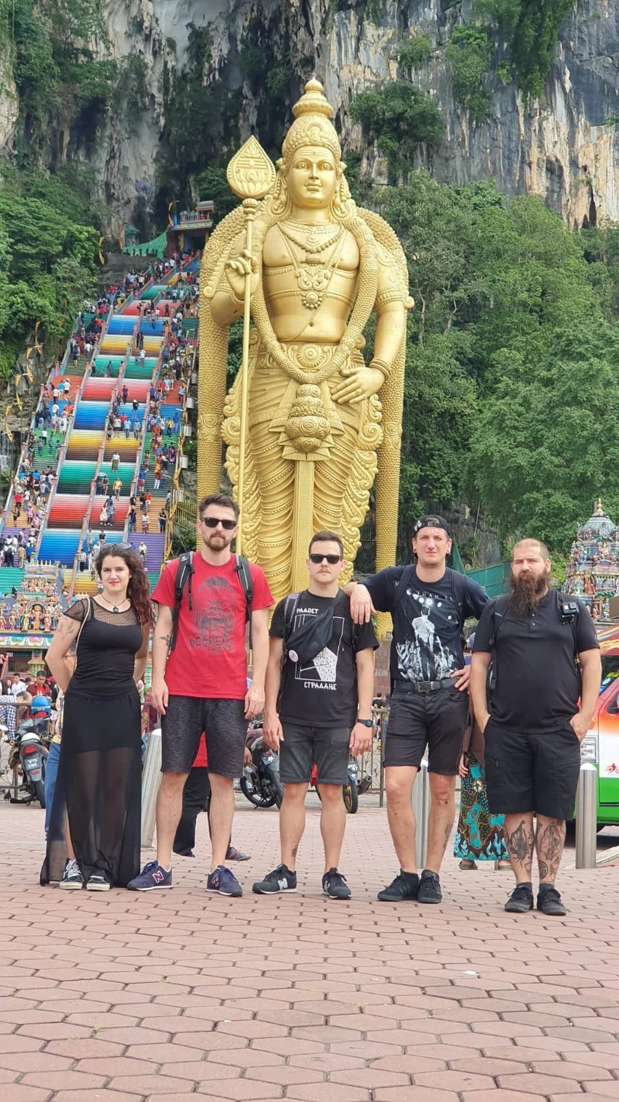 Group of five people standing in front of a large golden statue of Lord Murugan with colorful stairs leading up a hill behind them at Batu Caves in Malaysia.