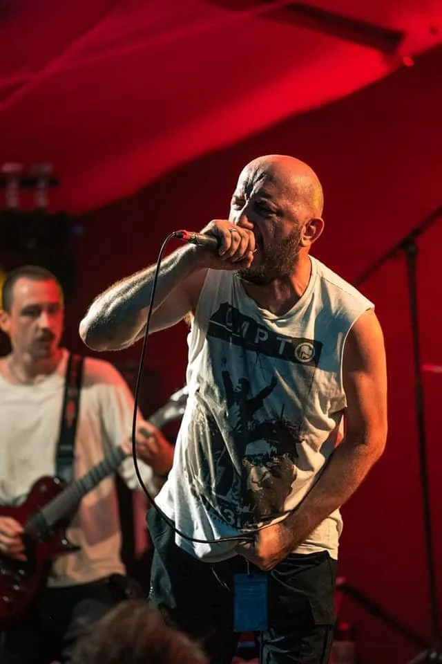 A vocalist passionately sings into a microphone during a live performance, with a guitarist playing in the background and red stage lighting.