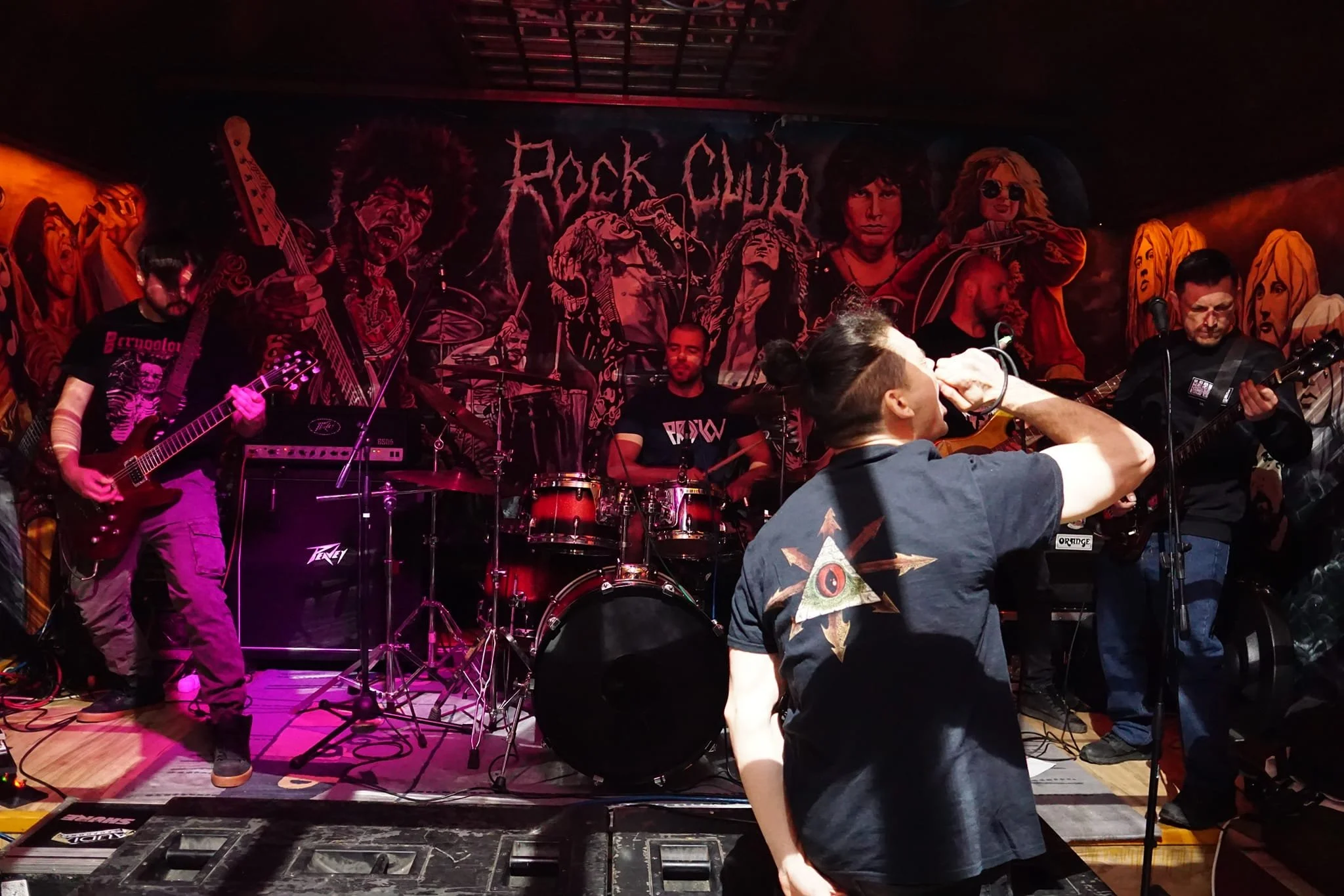 A music band performing on stage with a backdrop that reads 'Rock Outcast.' The band includes a guitarist, drummer, and vocalist, with audience members in the foreground.
