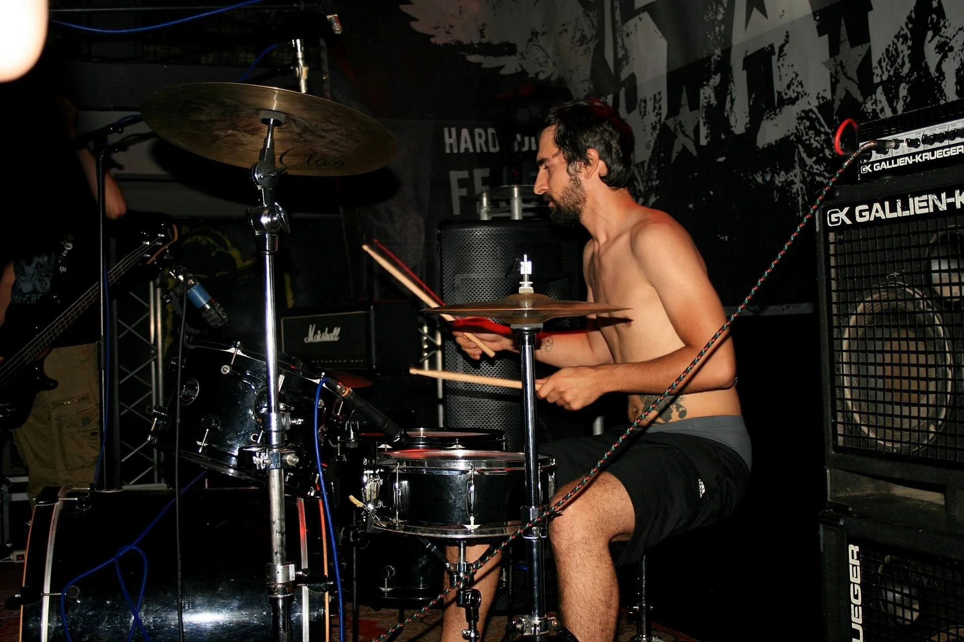 A shirtless man with dark hair and a beard playing drums with drumsticks on a drum set in a music venue, with various musical equipment and speakers around him.