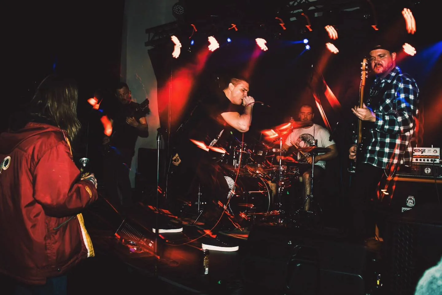 A live rock band performs on stage with a vocalist singing into a microphone, a guitarist playing, and a drummer in the background, illuminated by stage lights.