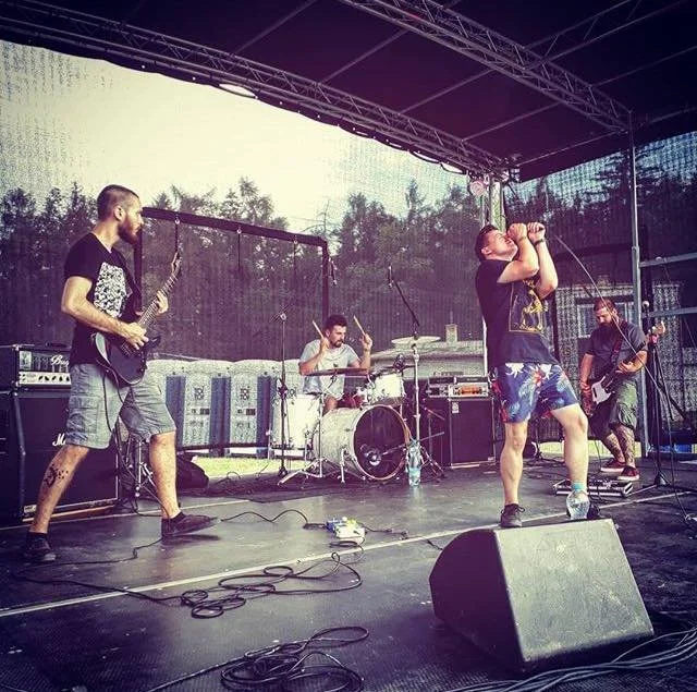 A four-member band performing on an outdoor stage with black backdrop and trees in the background, including a guitarist, drummer, bassist, and lead singer.