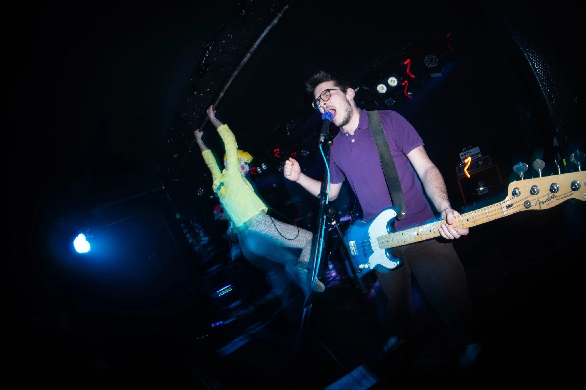 A male musician with glasses singing into a microphone and playing an electric bass guitar on stage in a dimly lit venue.