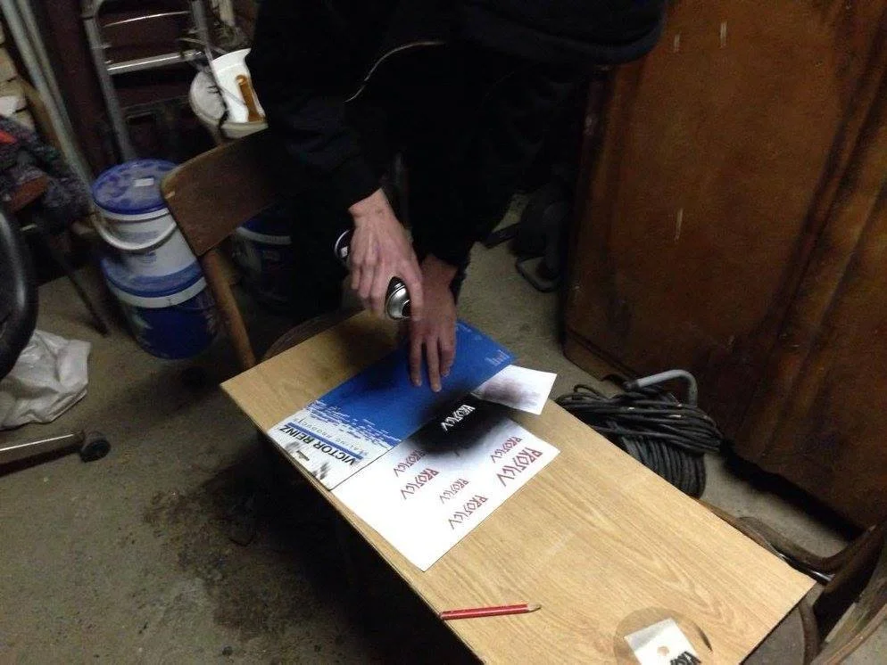 Person using a roller to apply black ink on a paper sign that says "PROSLOV" on a wooden table in a garage or workshop.
