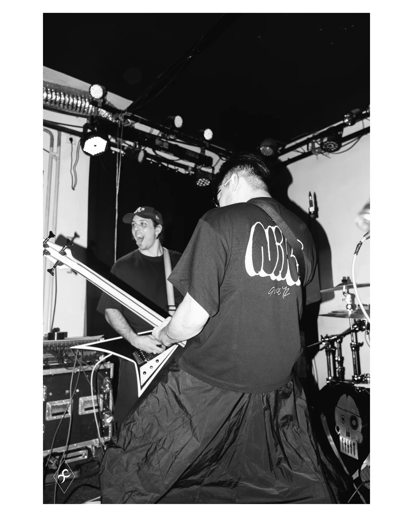 Two musicians performing on stage, one playing an electric guitar and the other singing, in a black-and-white photo with stage lights overhead.