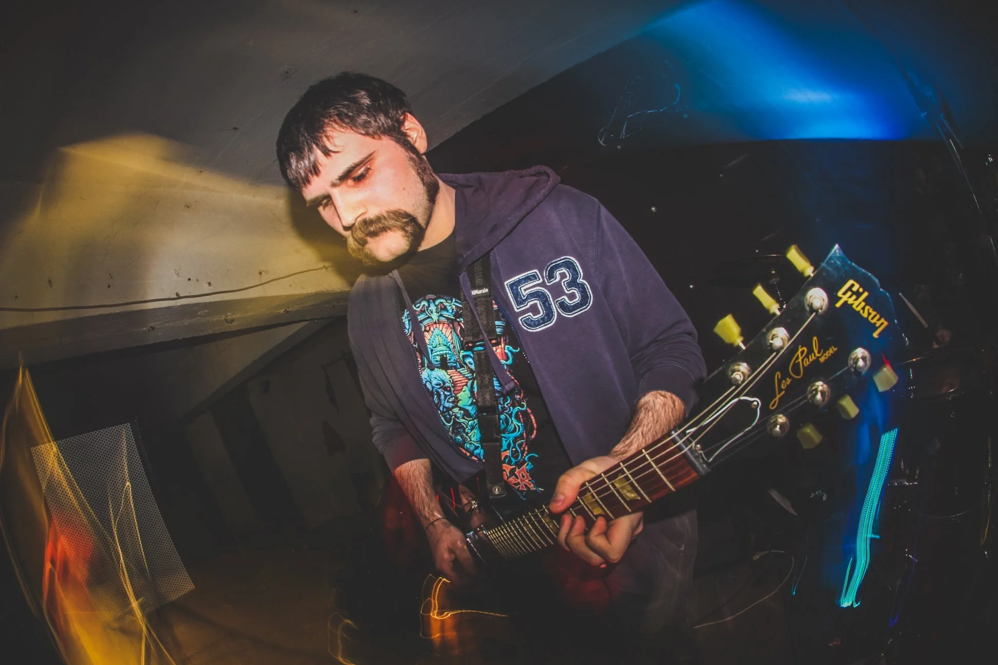 A man with dark hair and a beard, wearing a dark hoodie with the number 53, playing an electric guitar in a dimly lit room with colorful lighting.