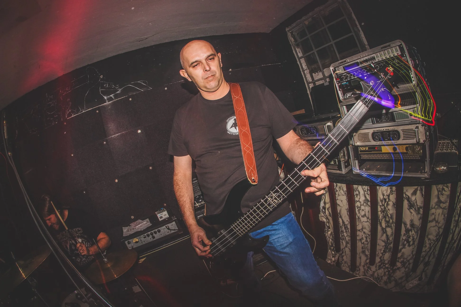 A man with a shaved head playing a black bass guitar in a dimly lit room with electronic music equipment behind him.