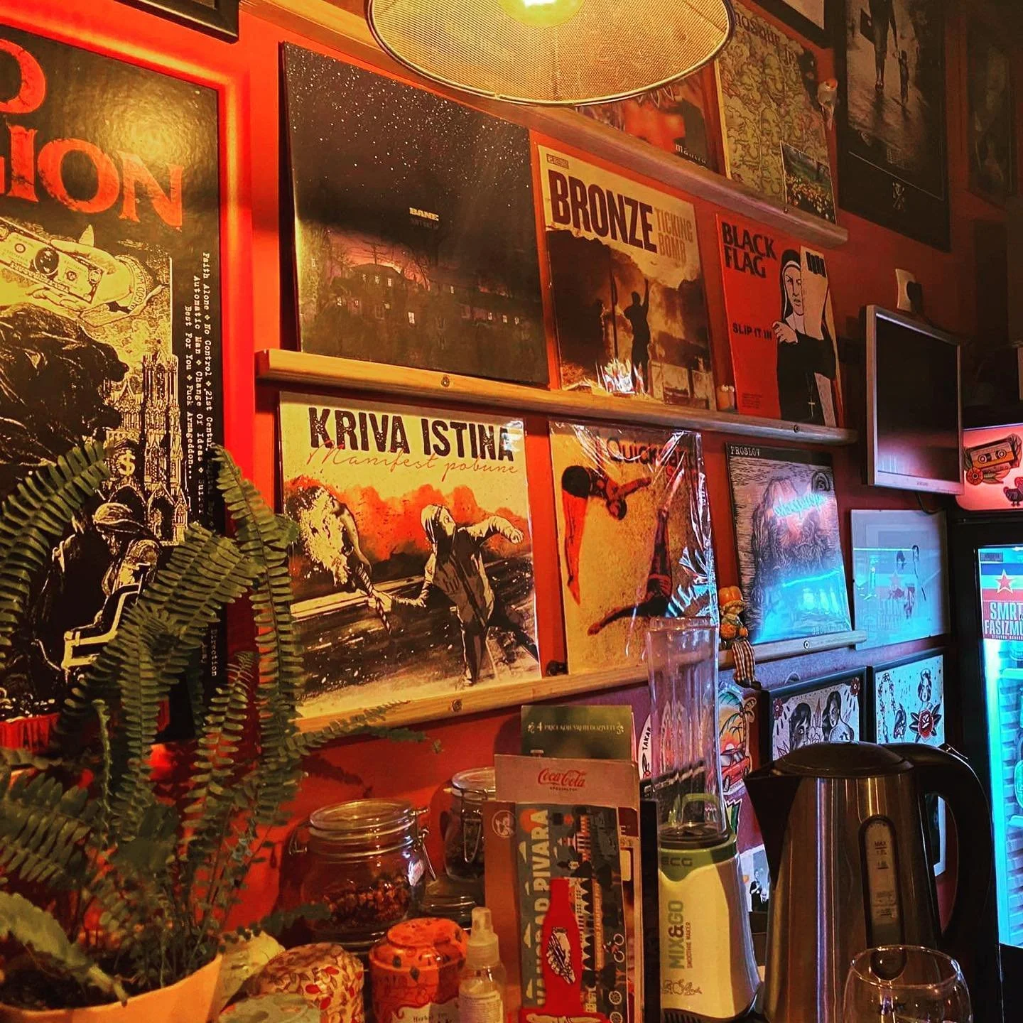 The image shows a wall decorated with various colorful posters, including album covers like 'Kriva Istina,' 'Bronze,' and 'Black Flag.' There are plants, jars, a glass of water, a vending machine, and a countertop with a metallic electric kettle and 