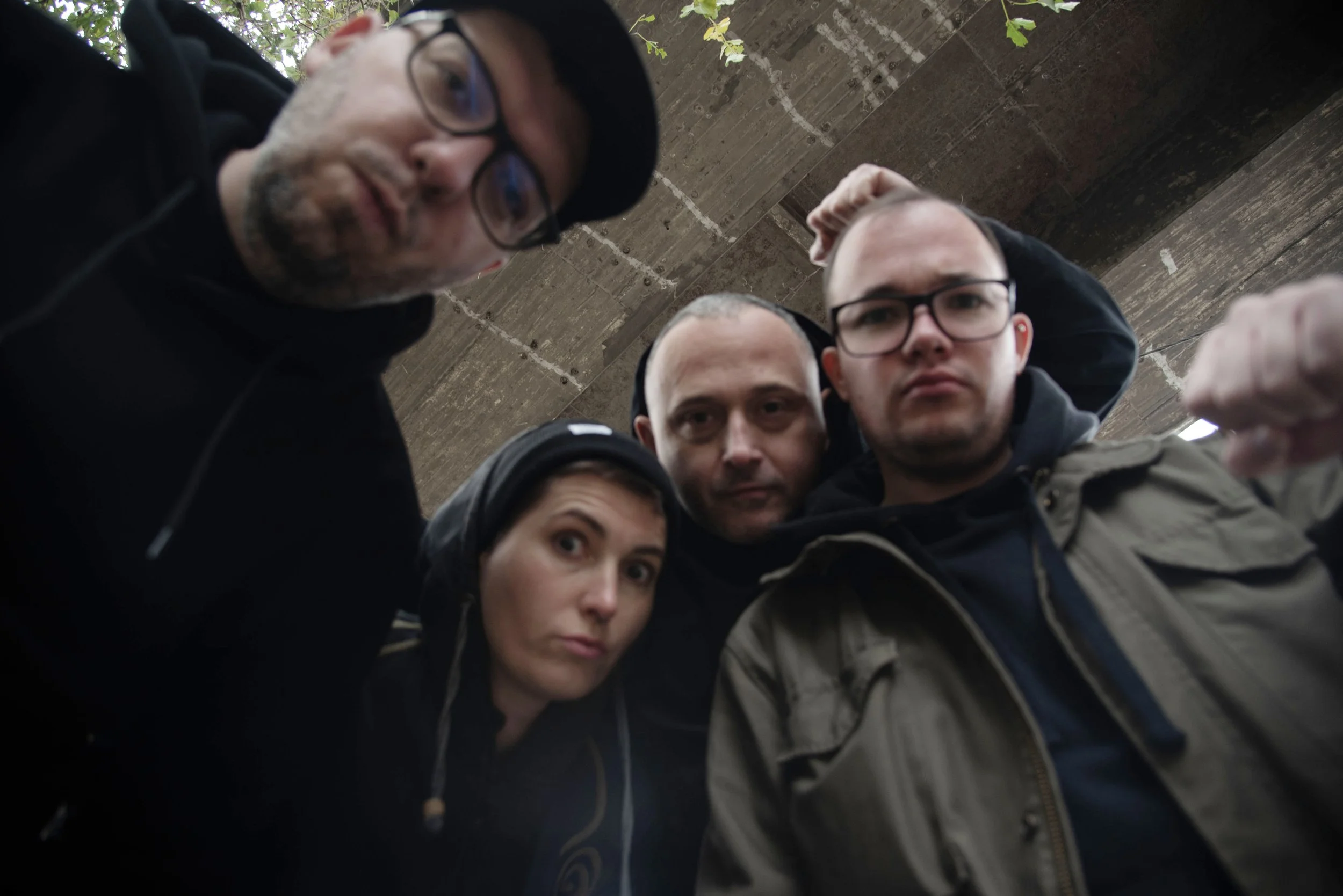 A group of five people looking down into the camera, taken from below, in an outdoor setting with a wooden ceiling and some green leaves visible.