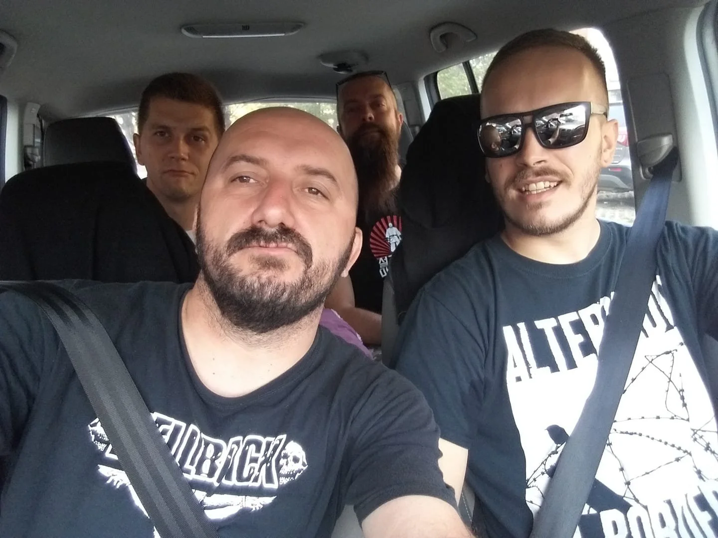 Four men sitting inside a car, with the driver wearing sunglasses and smiling, and the three passengers, two with neutral expressions and one with a beard, looking at the camera.