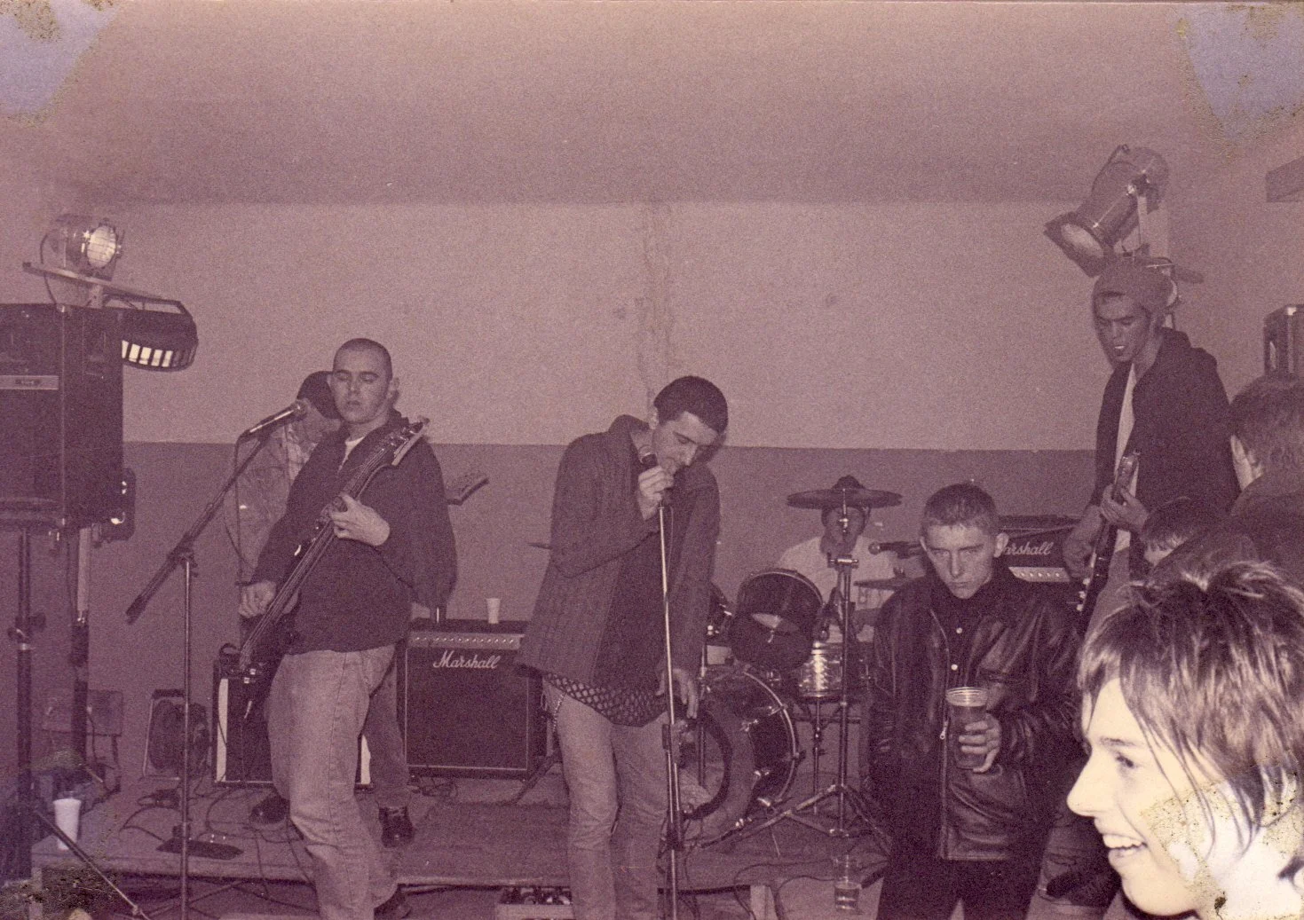 A group of young musicians performing on a small stage indoors, with posters or painted walls in the background, playing guitars, drums, and singing.