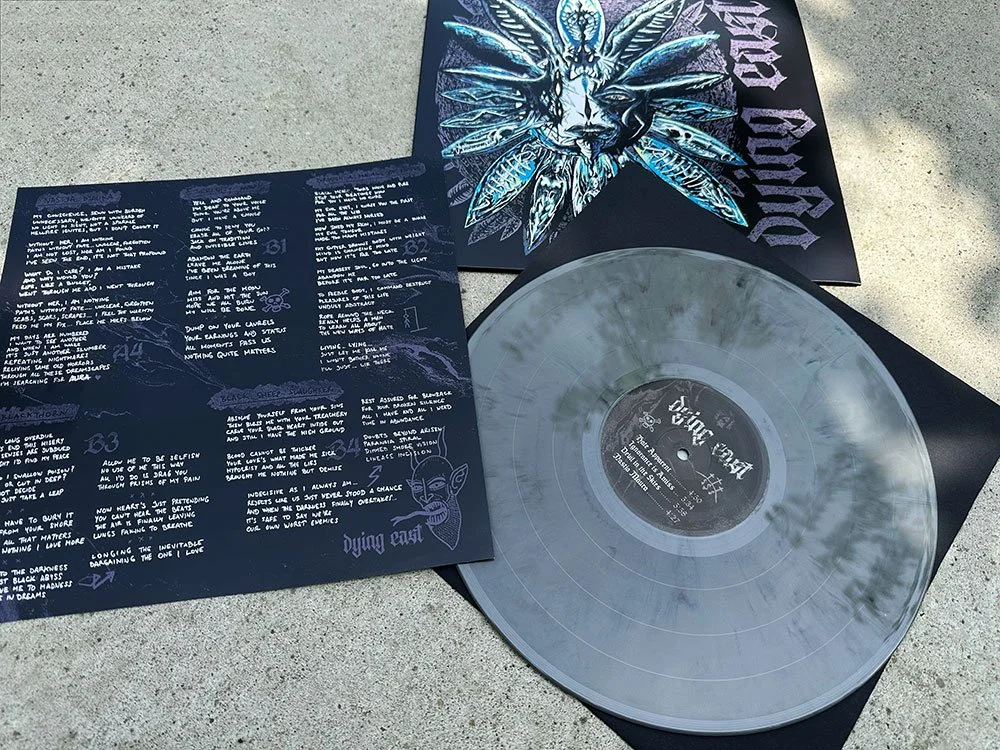 A vinyl record with a black and white marbled pattern on the record, placed on a black sleeve with purple and blue artwork. Next to the record, there is a lyric sheet with handwritten notes and drawings, and an album cover featuring a symmetrical blu
