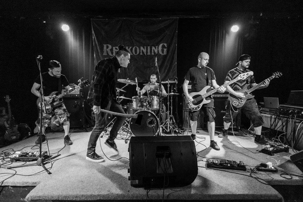 A band of five musicians performing on stage with guitars, drums, and microphones, under a banner that says ALL EXCEPT ONE.