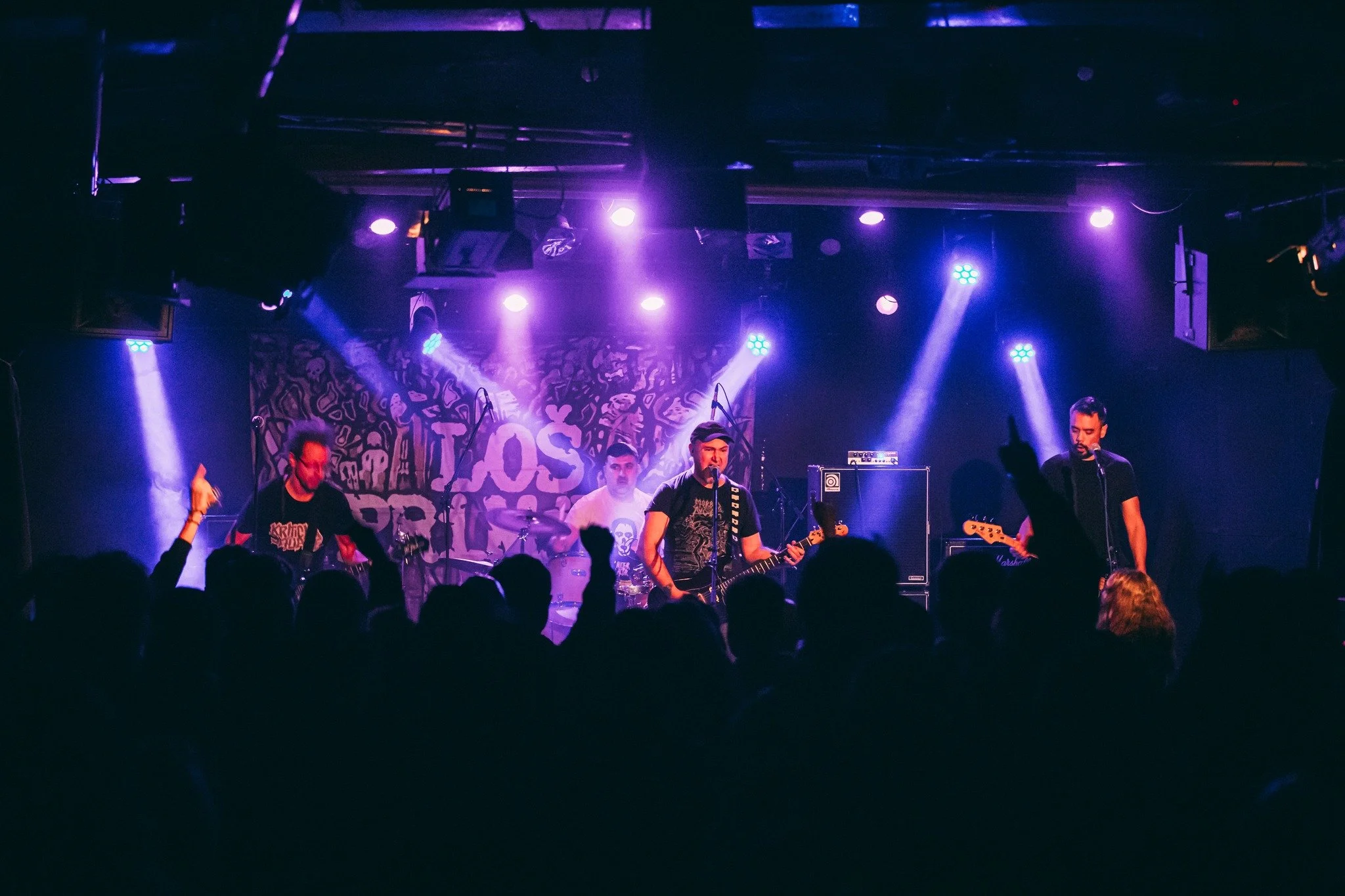 A live music performance on stage with multiple band members playing instruments under purple stage lights, and a backdrop with artwork and text.