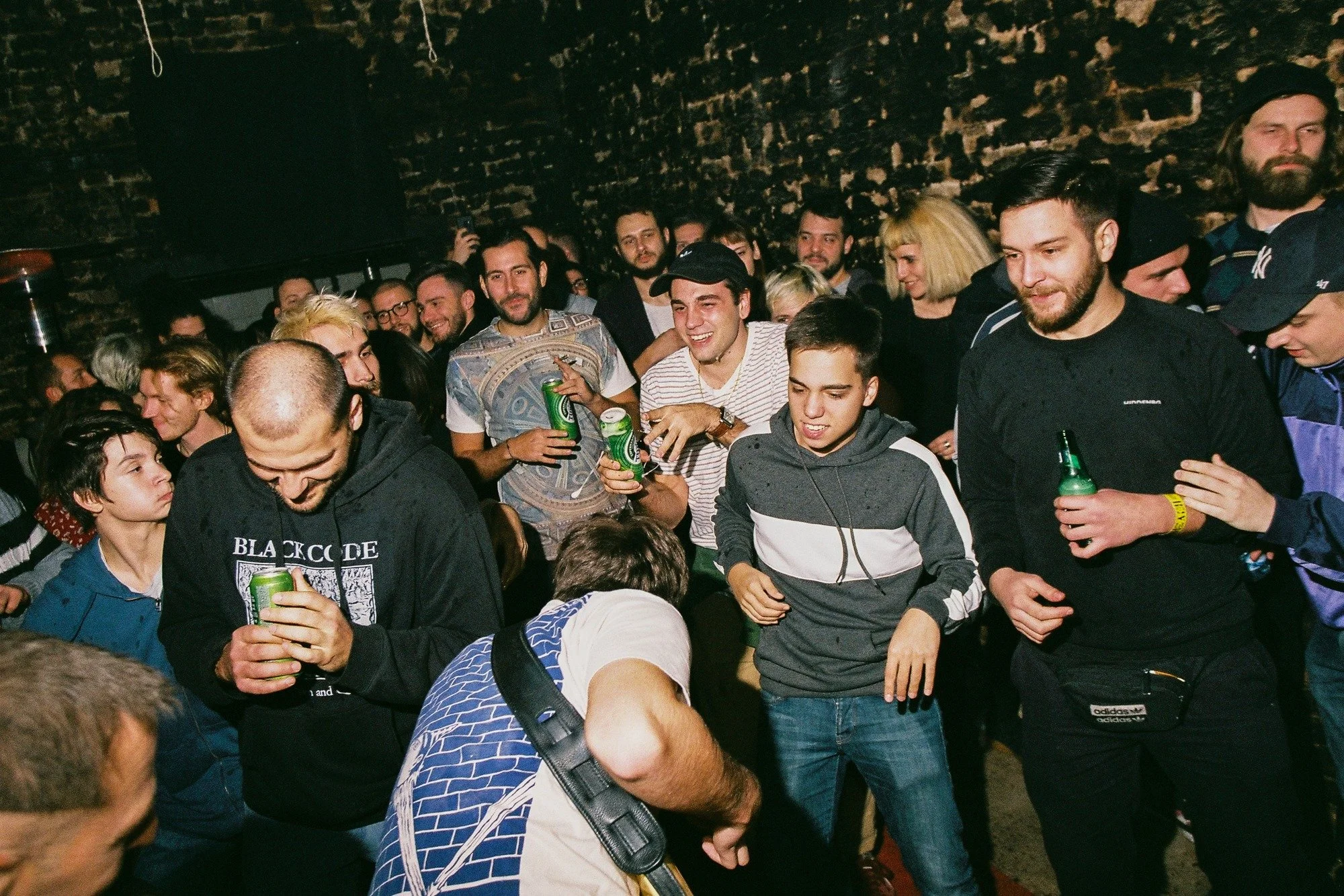 A crowded indoor party with young adults socializing and drinking beer, in front of a dark brick wall.