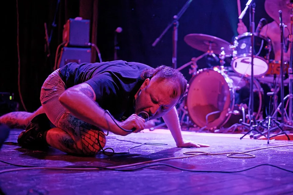 A performer crouches on stage, singing into a microphone, with a drummer playing in the background.