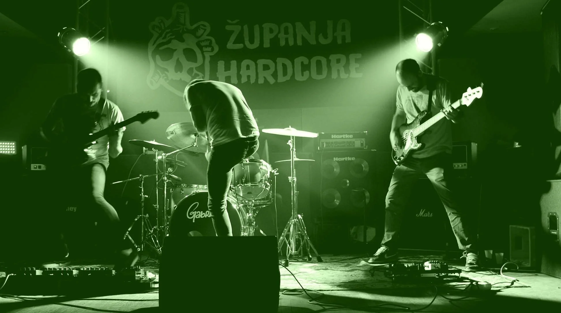 A band performing live on stage with drums and electric guitars, with a backdrop that says 'ZUPANJA HARDCORE'.