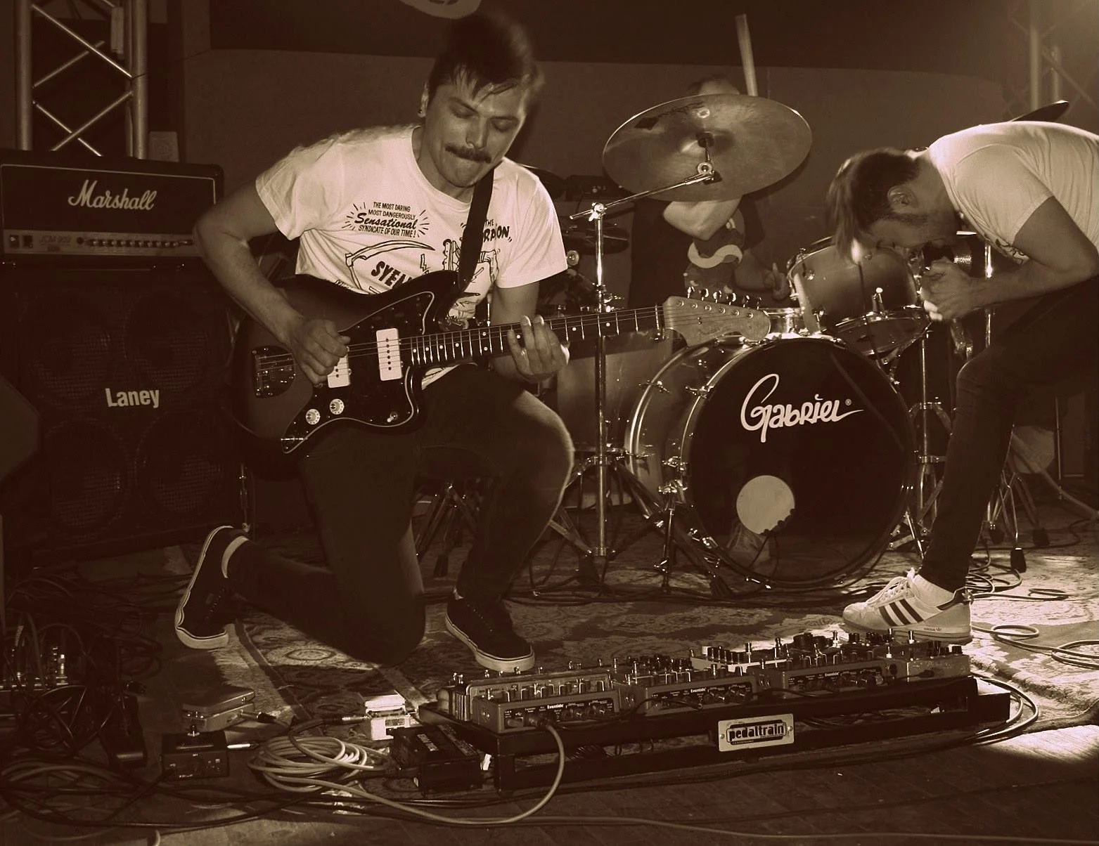 Two musicians performing with electric guitars and a drum set on stage, with musical equipment and an amplifier visible.
