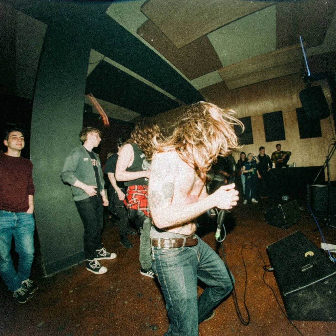 A man with long curly hair singing or speaking into a microphone at a crowded indoor event or concert. Several people are standing in the background against a wall in a dimly lit room with a wooden ceiling while band SUPLEXX is playing.