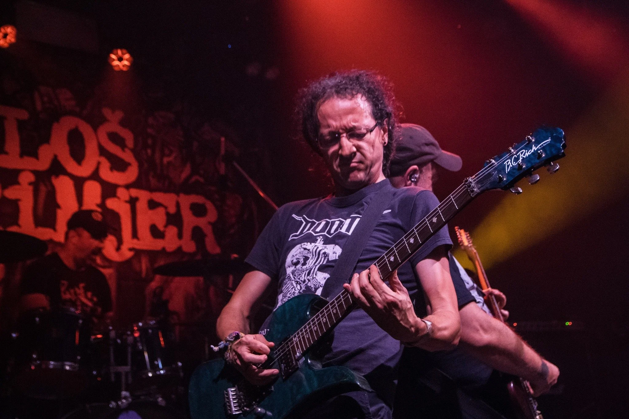 A guitarist performing on stage with a dark background and red lighting, featuring a drummer in the background and a partial view of another band member.