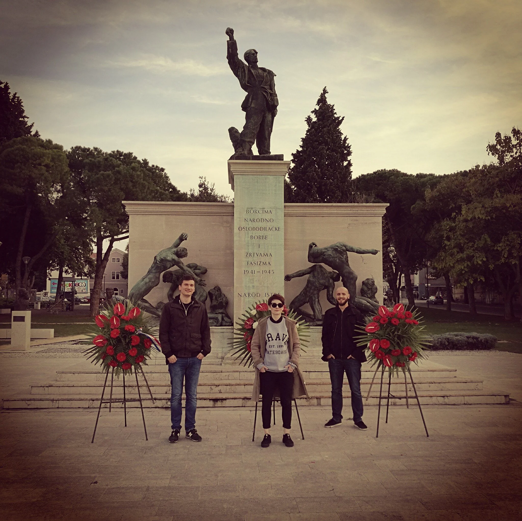 A monument with a statue of a man holding a clenched fist, surrounded by bronze sculptures of people in various poses, with three people standing in front of it holding flower arrangements.