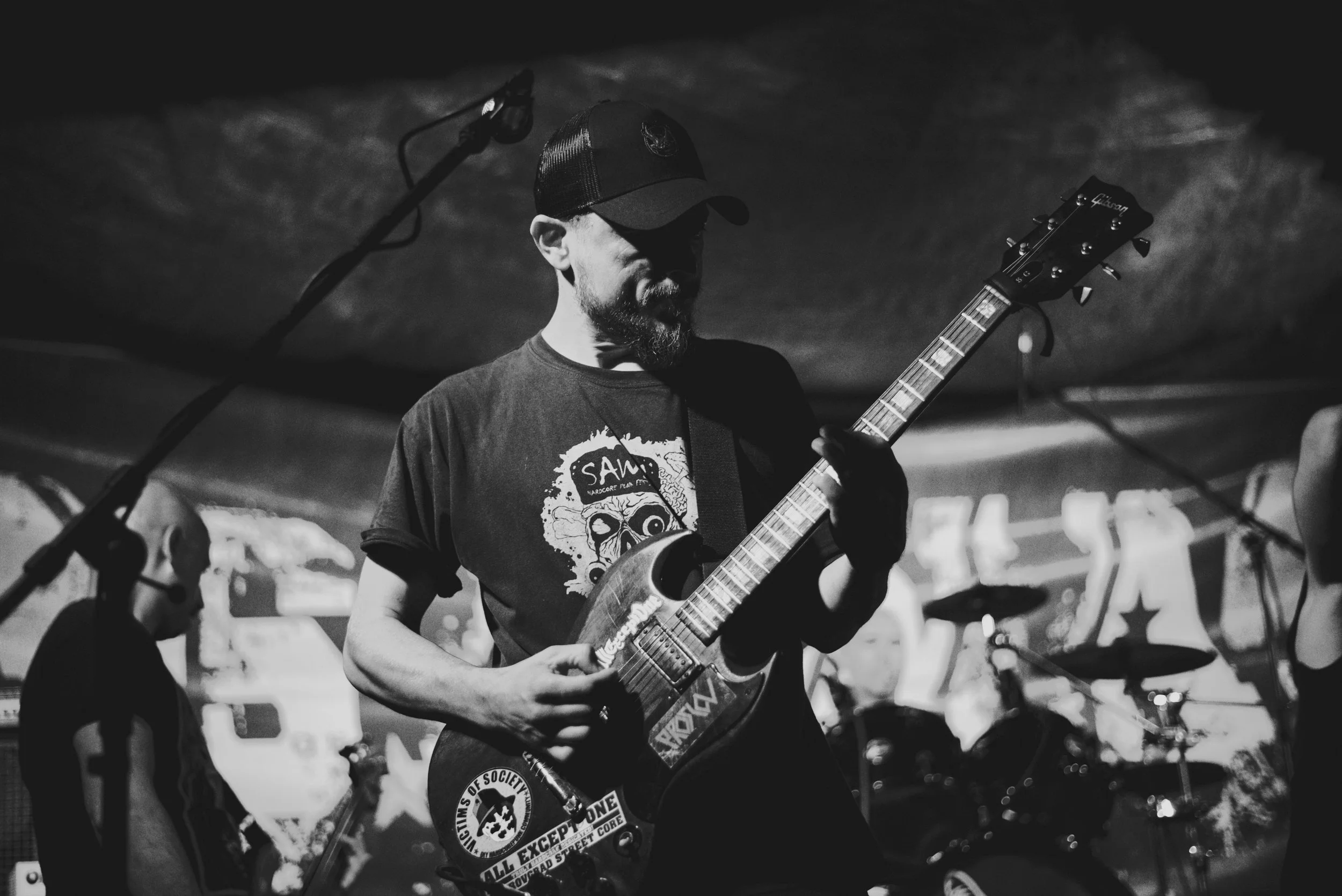 A man wearing a baseball cap and a graphic t-shirt playing electric guitar on stage, with a microphone in front of him and other musicians in the background.