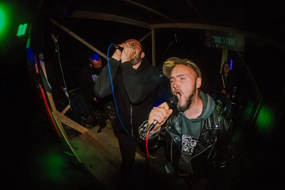 Two men passionately singing into microphones at a live music performance. One with a bald head, black hoodie, and the other with a cap and a leather jacket, on a small stage with dim lighting.