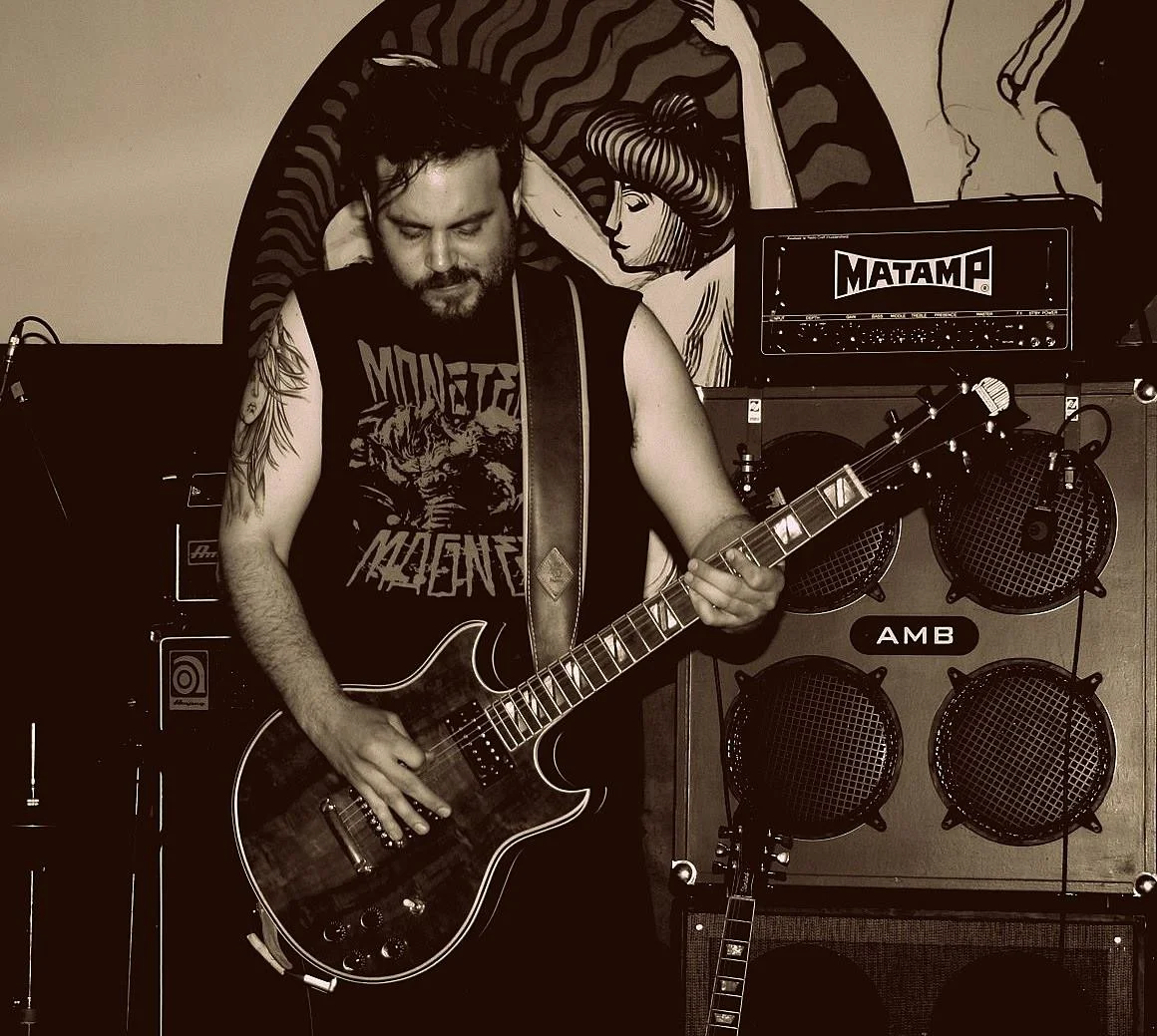A man playing an electric guitar on stage, wearing a Metallica t-shirt, with tattoos on his arms, in front of a wall with artwork of a woman and a wall of speakers and guitar amps.