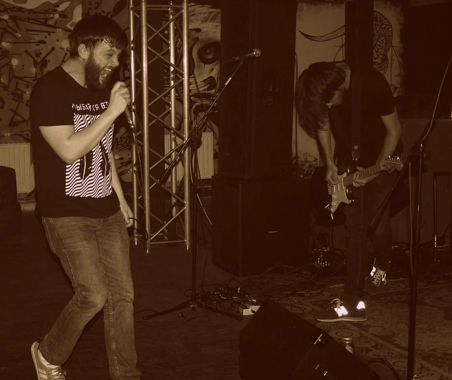 A singer with a beard, wearing a black t-shirt and jeans, performs with a microphone on stage. A guitarist with long hair, dressed in black, plays an electric guitar nearby. The stage background features graffiti-style artwork.