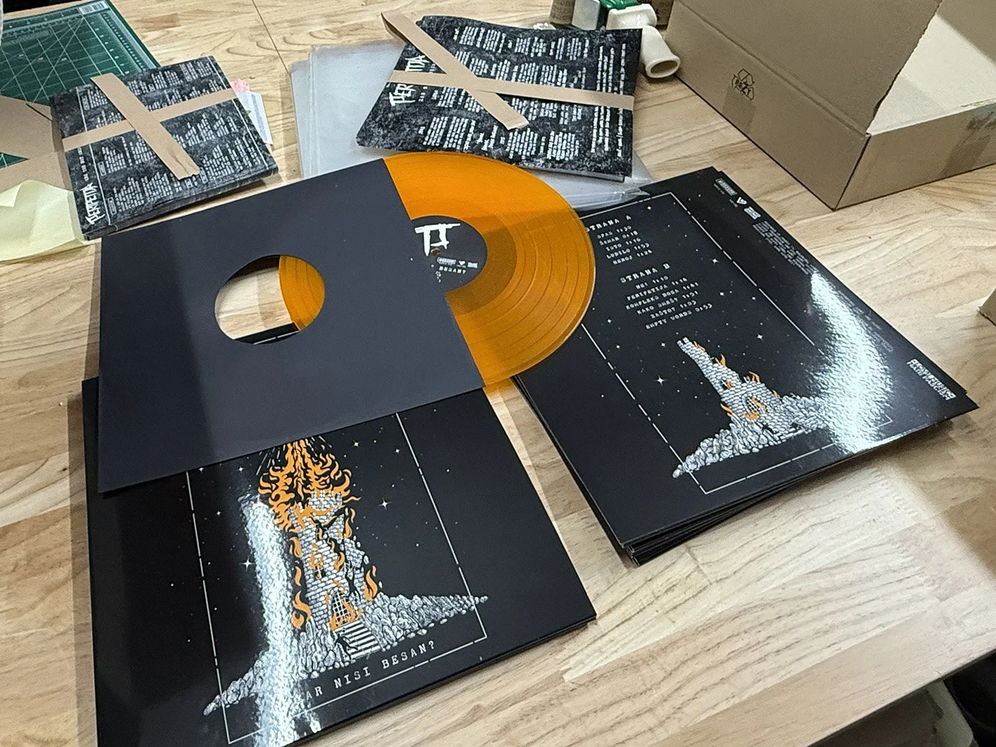A vinyl record partially removed from black sleeves with artwork of a rocketship and flames, on a wooden table with scattered items including paper, tape, and boxes.