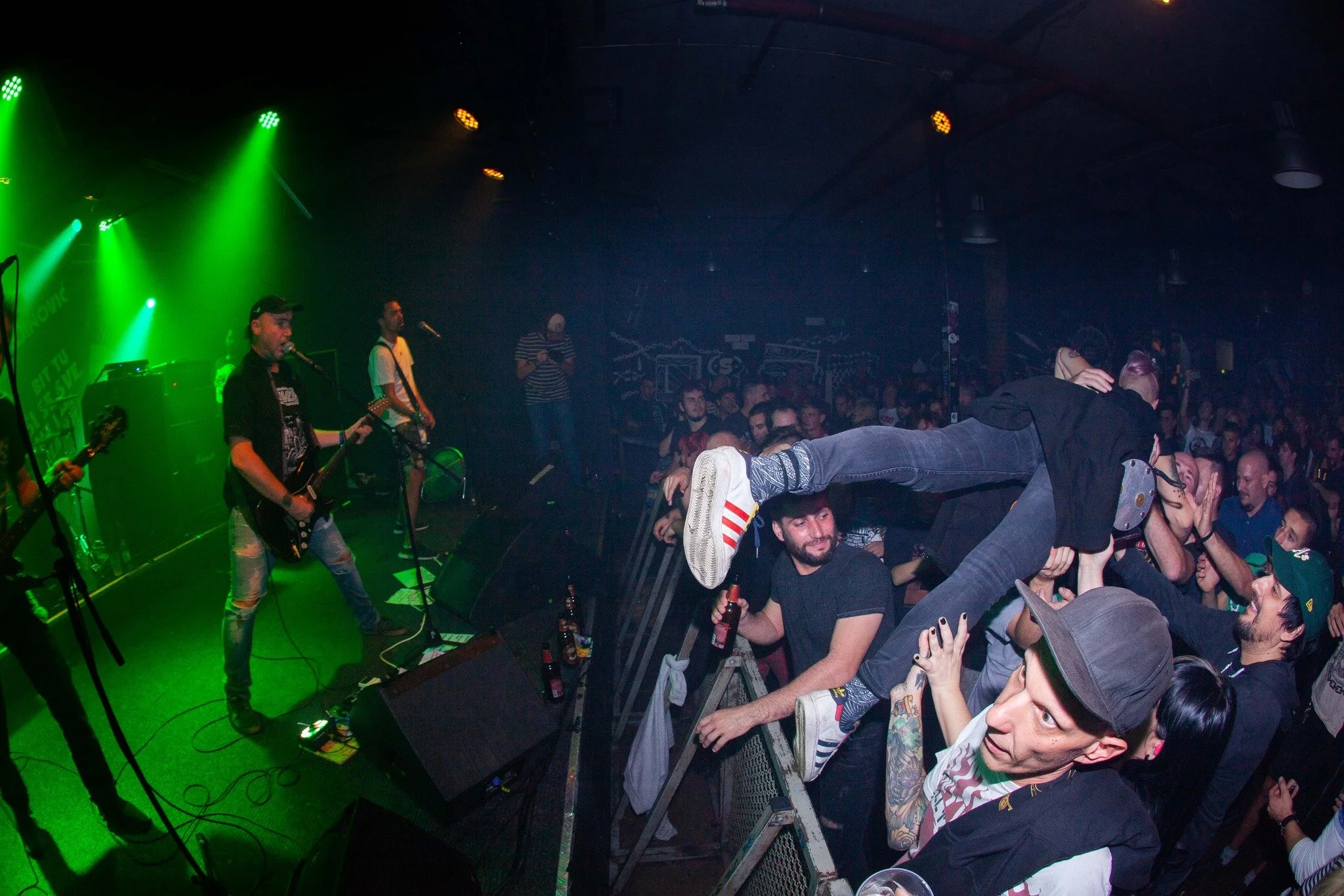 A live music concert with band members on stage performing under green lighting, and a crowd of people in the audience, some crowd surfing and enjoying the show.