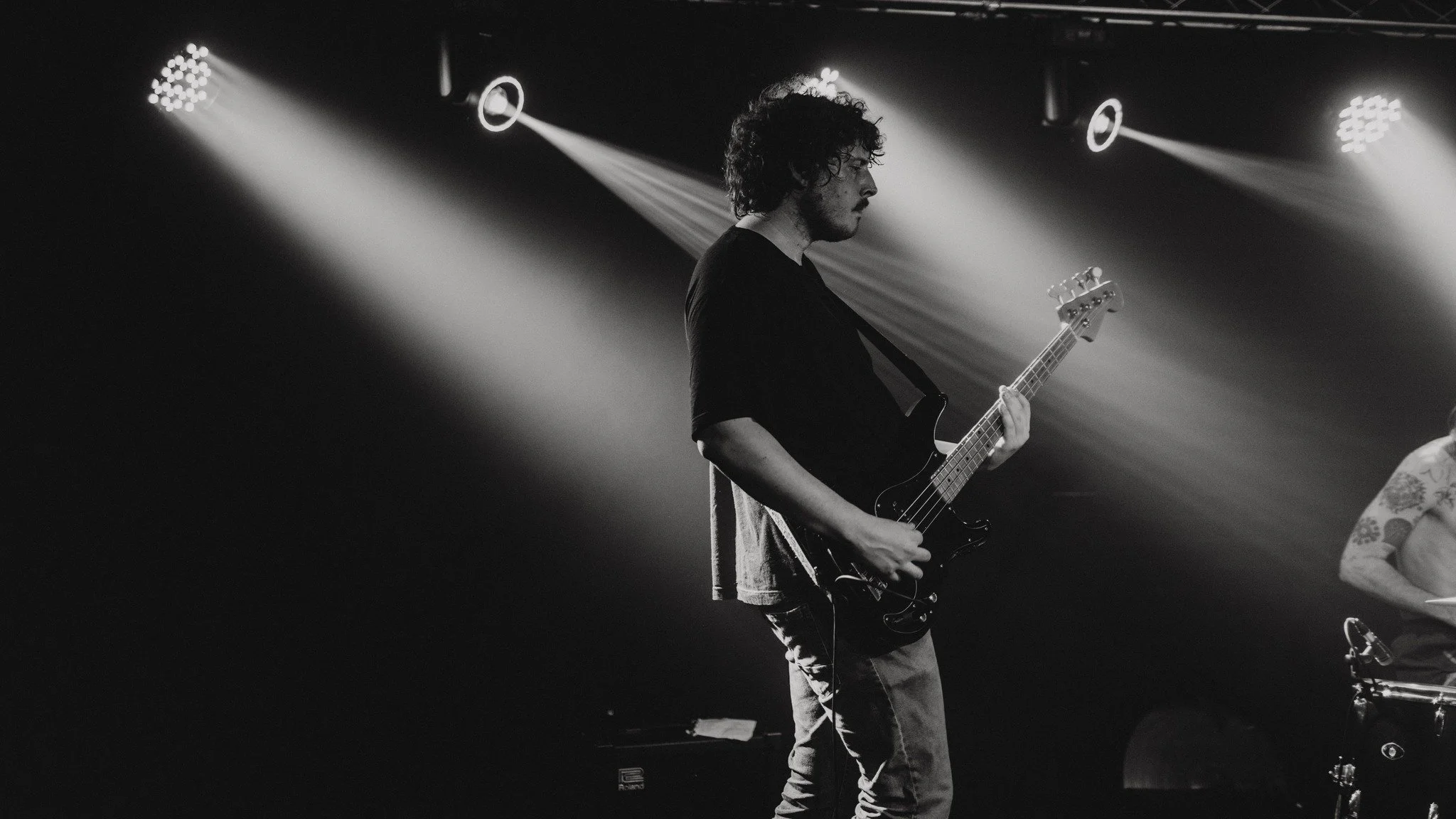 A musician with curly hair playing an electric guitar on stage under bright spotlights, in black and white.
