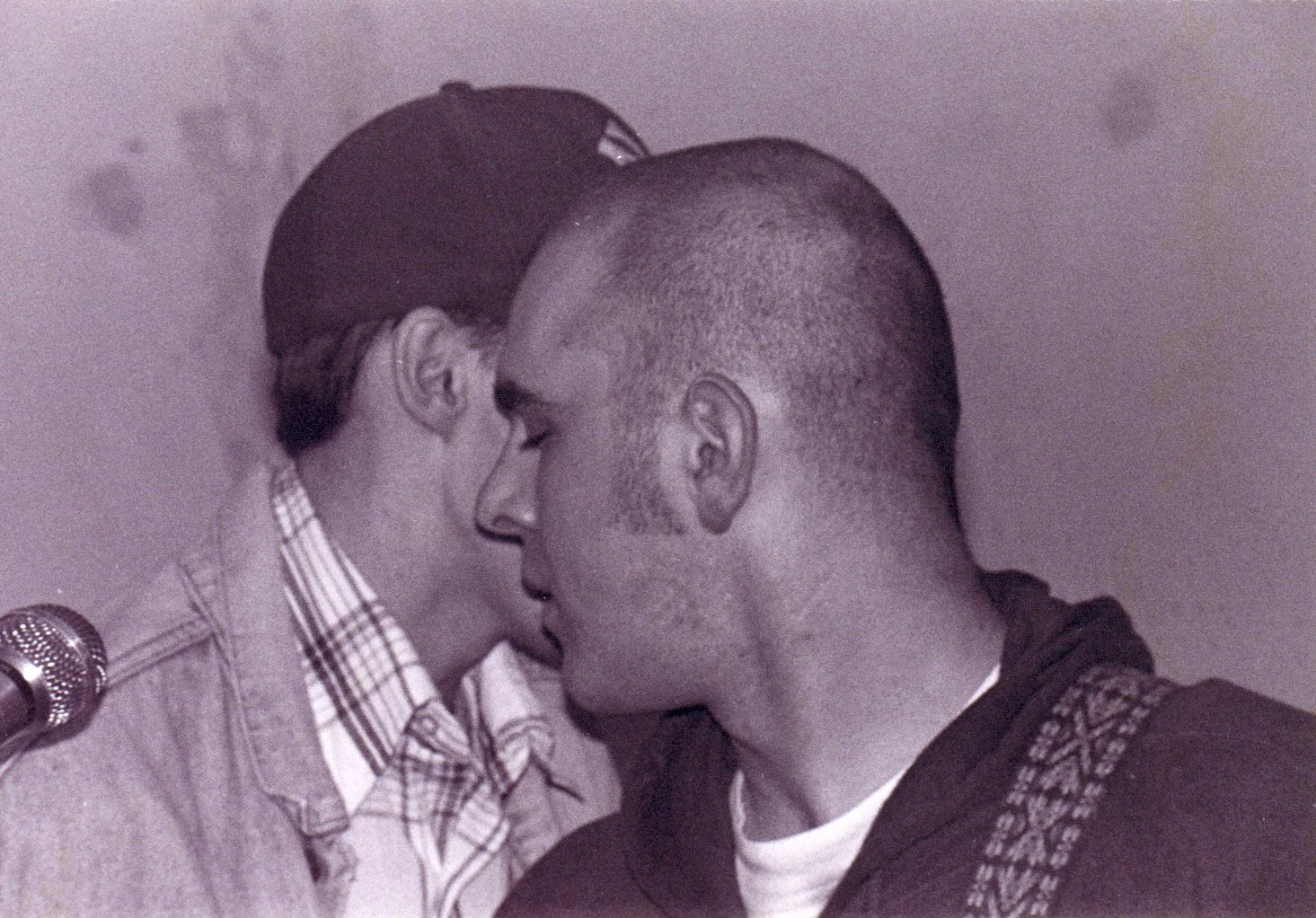 Two men are close together with foreheads touching, eyes closed, in a gesture of intimacy or comfort. One man has a shaved head and is wearing a hoodie, the other man has dark hair and is wearing a checked shirt and glasses.