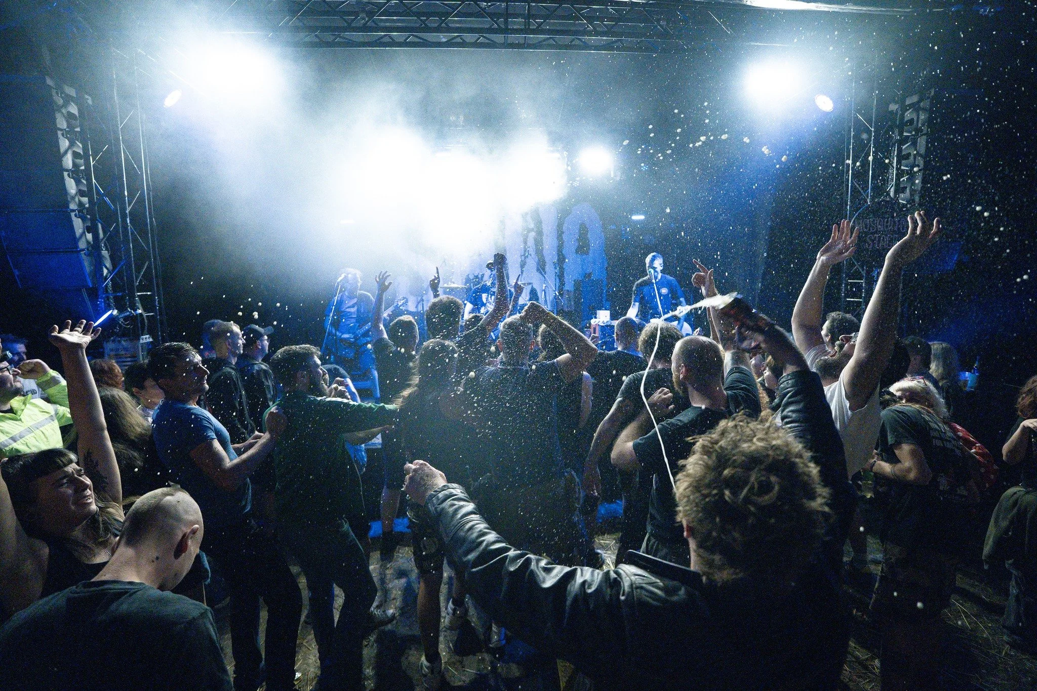 Concert stage with bright lights, a band performing, and a crowd dancing and enjoying themselves, with some people with hands raised, in a lively music event.