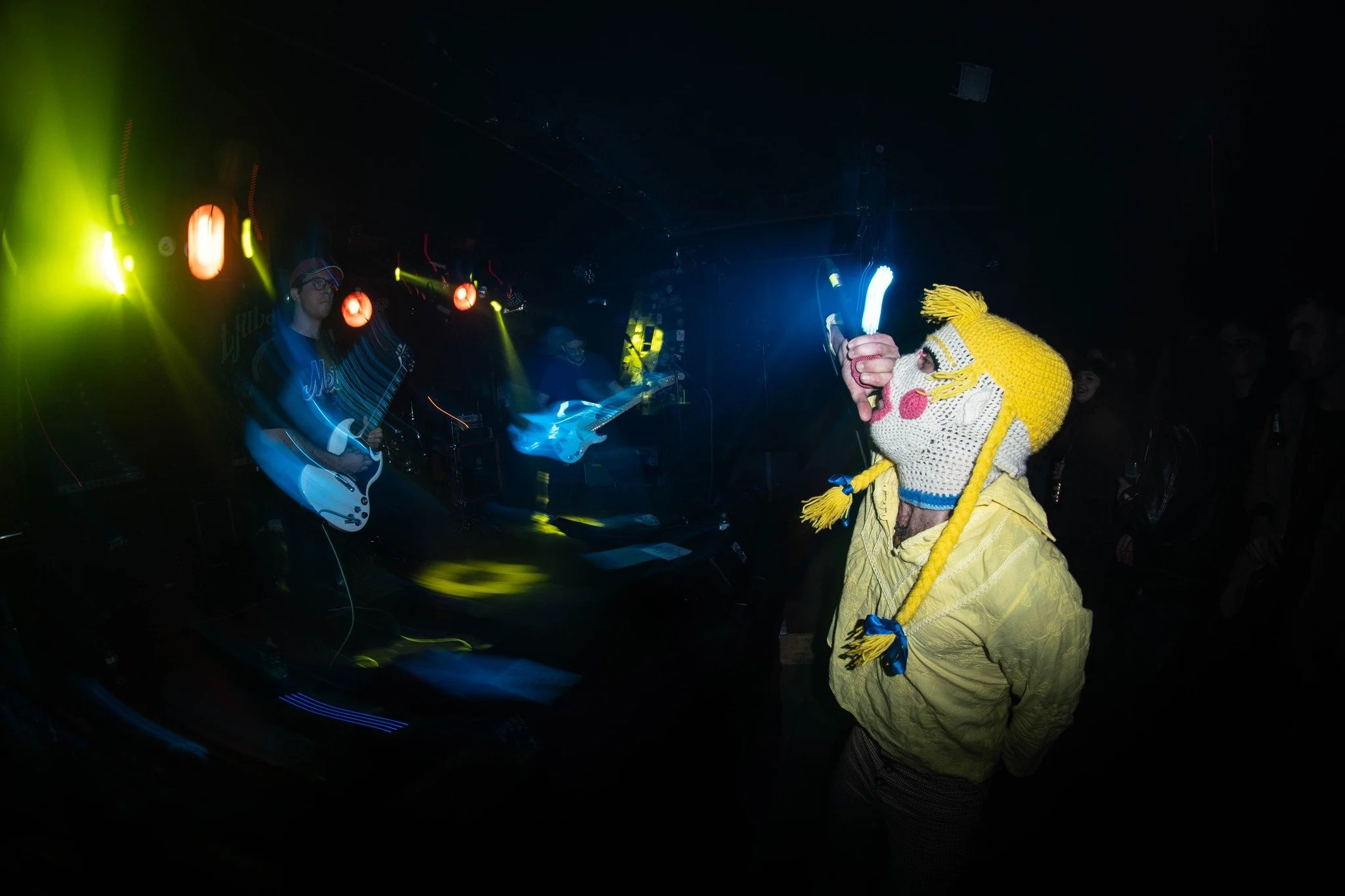 A person wearing a yellow coat and a knitted hat with braided tassels is holding a glow stick at a live music concert. The stage features colorful lighting and musicians playing guitars.