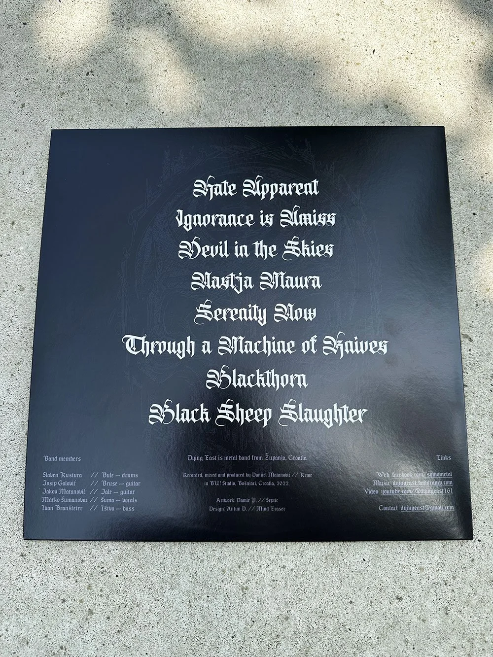 Black album cover with gothic white text listing song titles and band members, on a concrete ground.