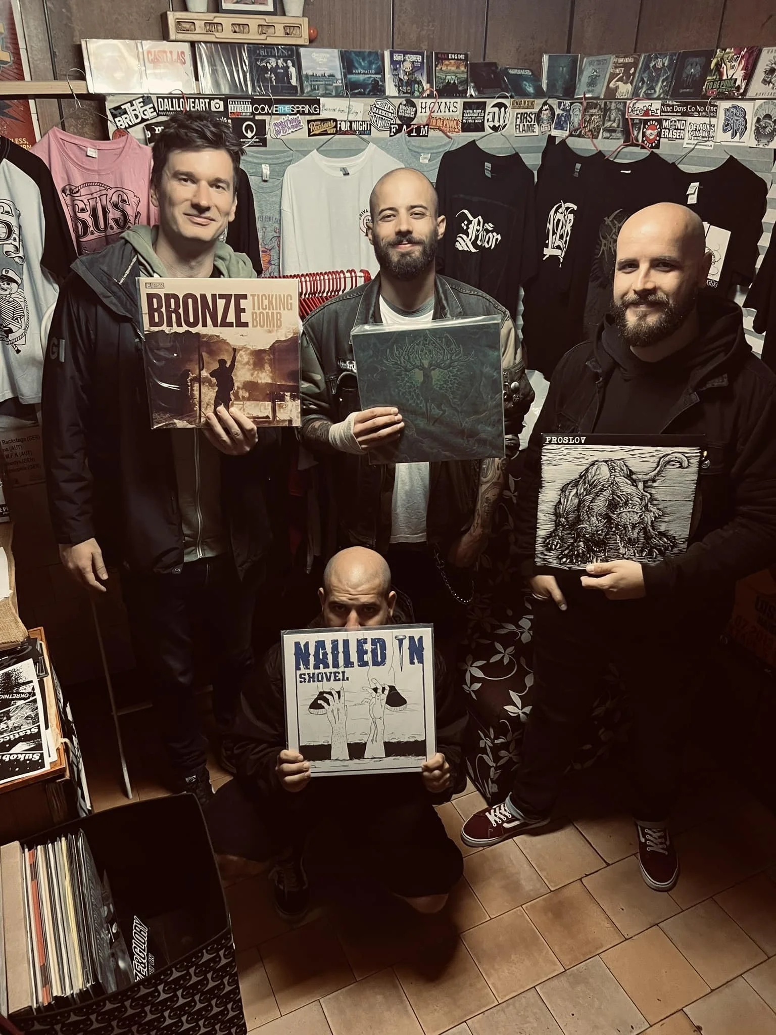 Three men standing in a record store, holding vinyl records, with rack of T-shirts and posters behind them.