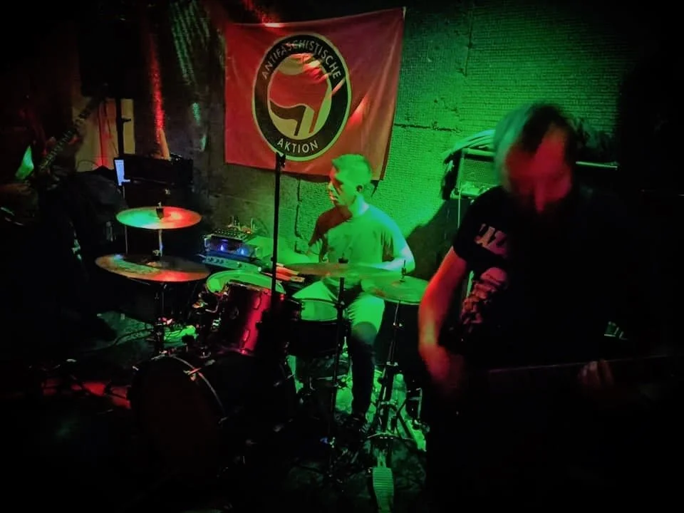 Two musicians performing on stage with green and red lighting, one playing drums and the other playing guitar, with a banner in the background that reads 'Antifaschistische Aktion'.