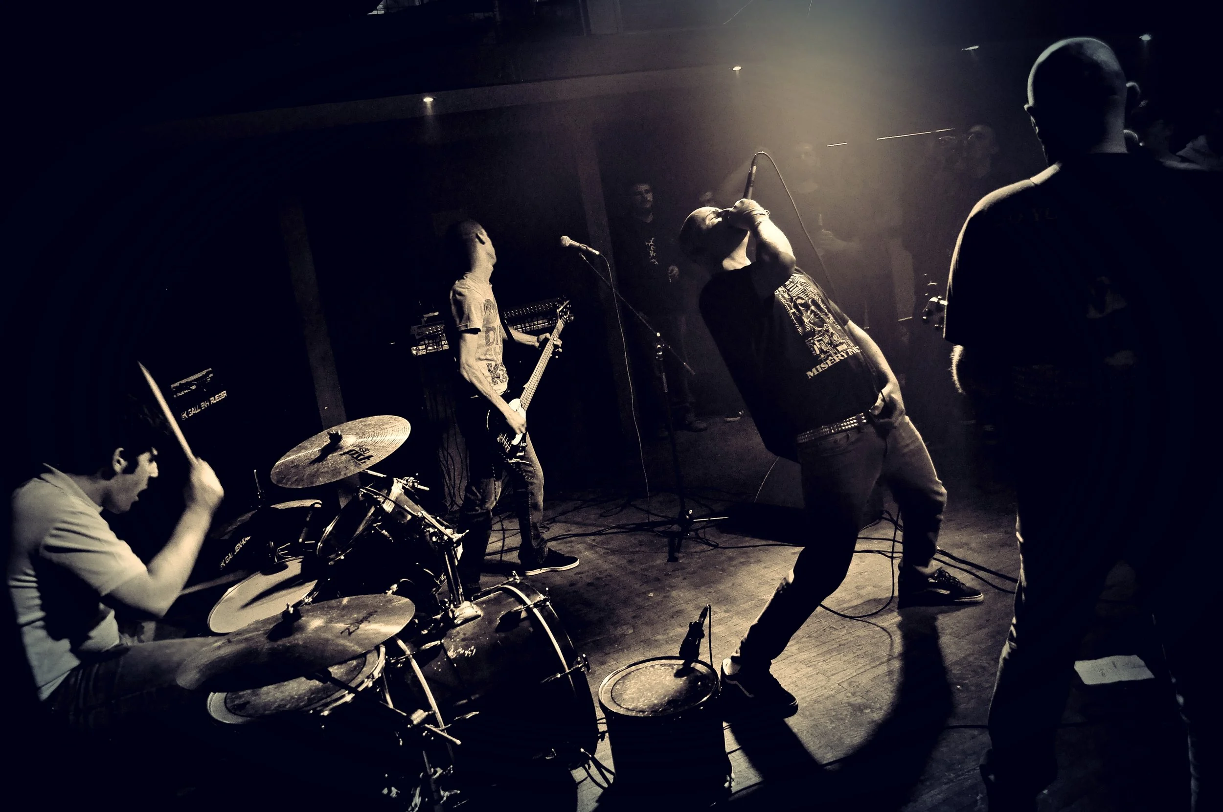 A band performs on stage in a dimly lit room. The band includes a drummer, a guitarist, a vocalist, and a person playing bass. The scene has a dark and moody atmosphere with spotlights highlighting the performers.