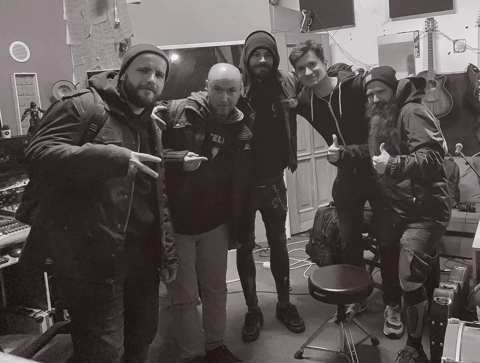 A group of six men posing together in a music studio. They are wearing casual outdoor clothing, some with hats and beanies, and are standing in front of musical equipment and guitars on the wall.