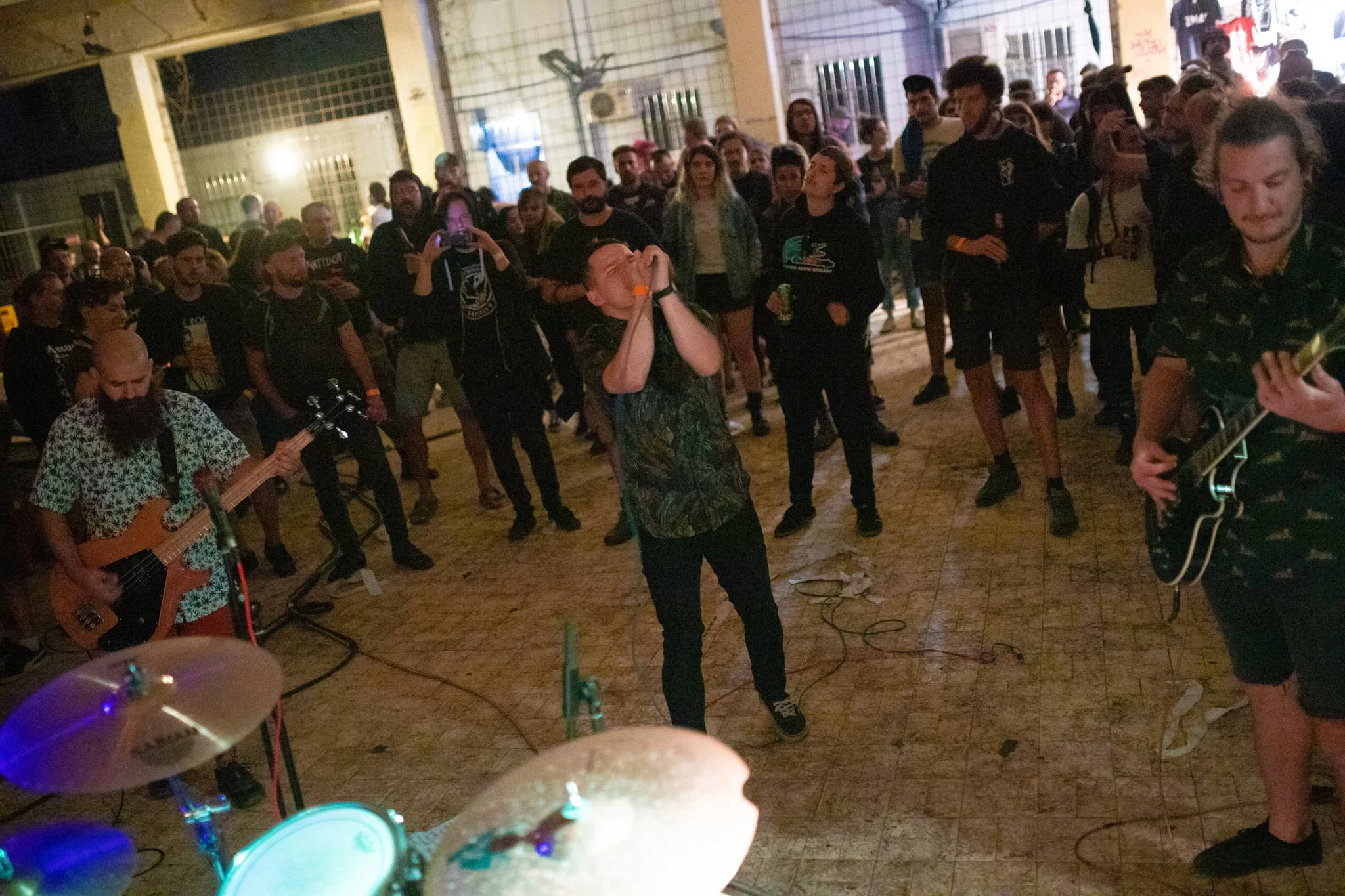 A group of people gathered indoors at night, with a man singing into a microphone at the center. Some are playing musical instruments, and the background shows a cage-like structure and graffiti on the wall.