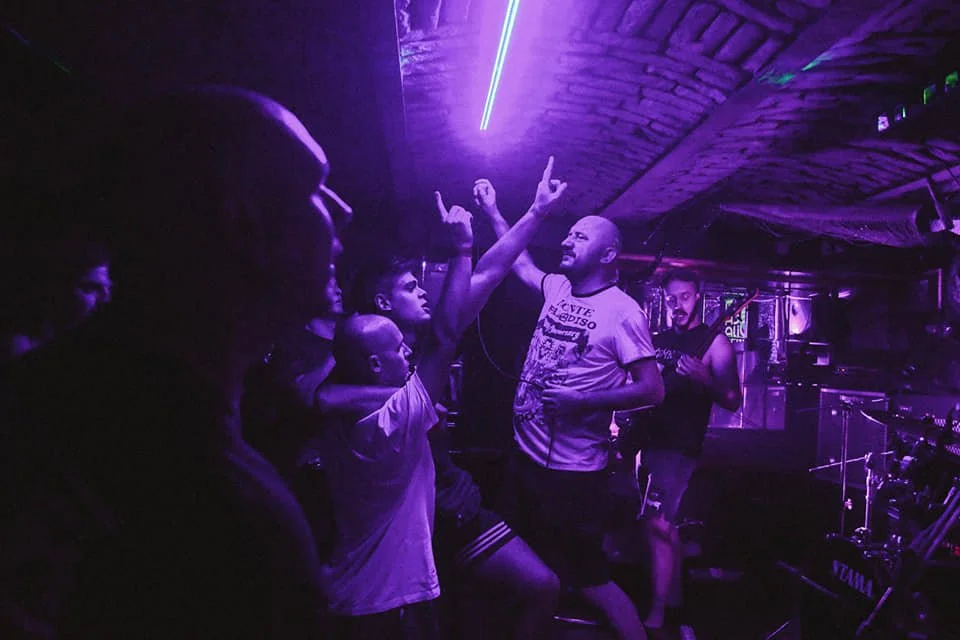 People dancing and enjoying music in a dimly lit club with purple lighting, with one person raising their hands and smiling.