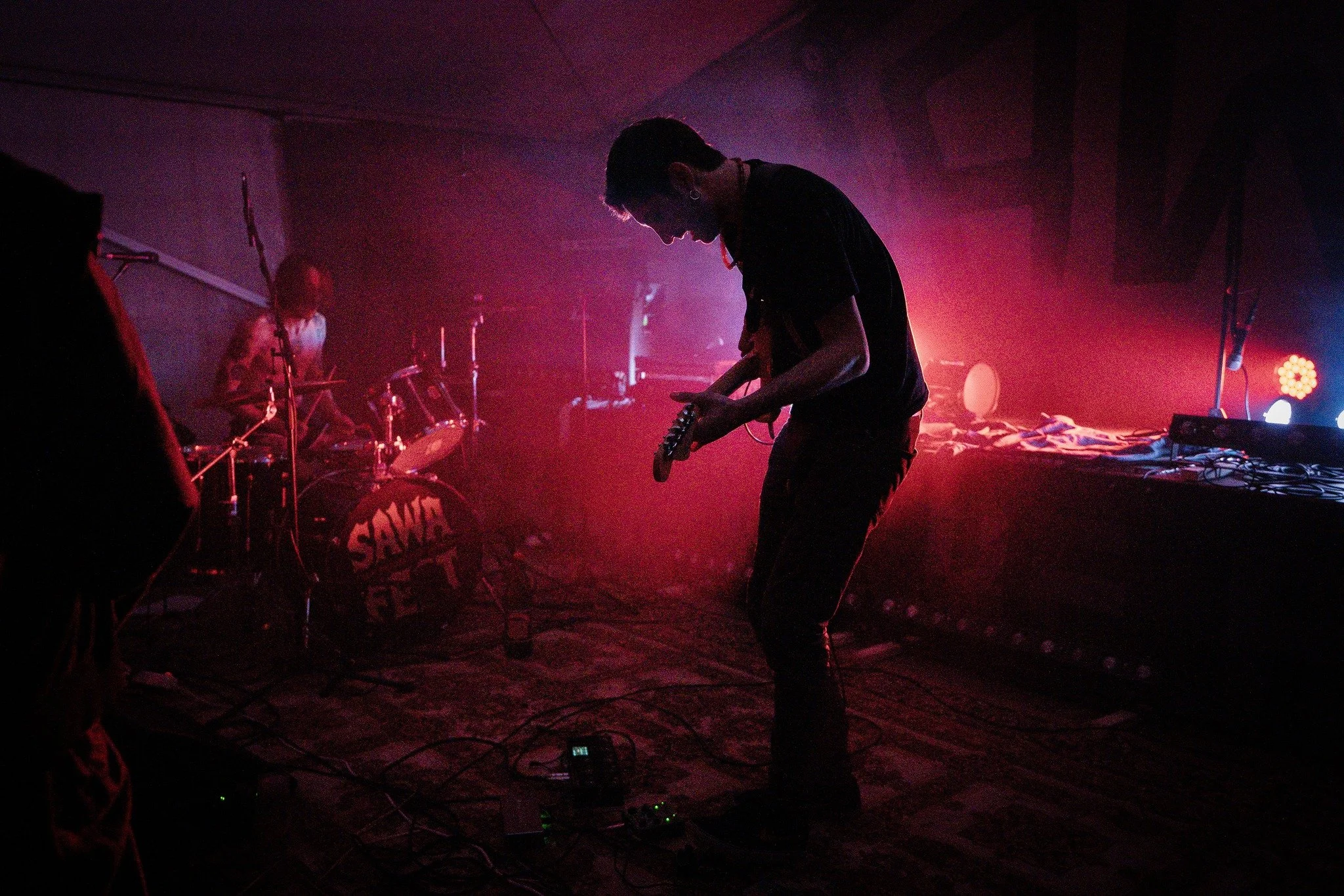 Dark scene of a live music performance with two musicians, one holding a guitar and the other playing drums, illuminated by purple and pink stage lighting.