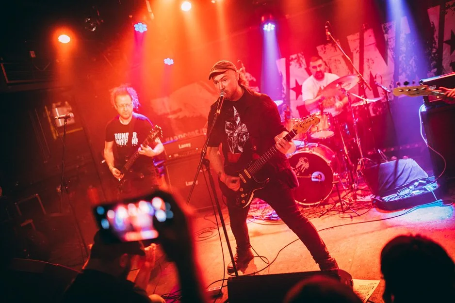 A band performing live on stage with colorful lighting, including a guitarist and a drummer, in front of an audience.