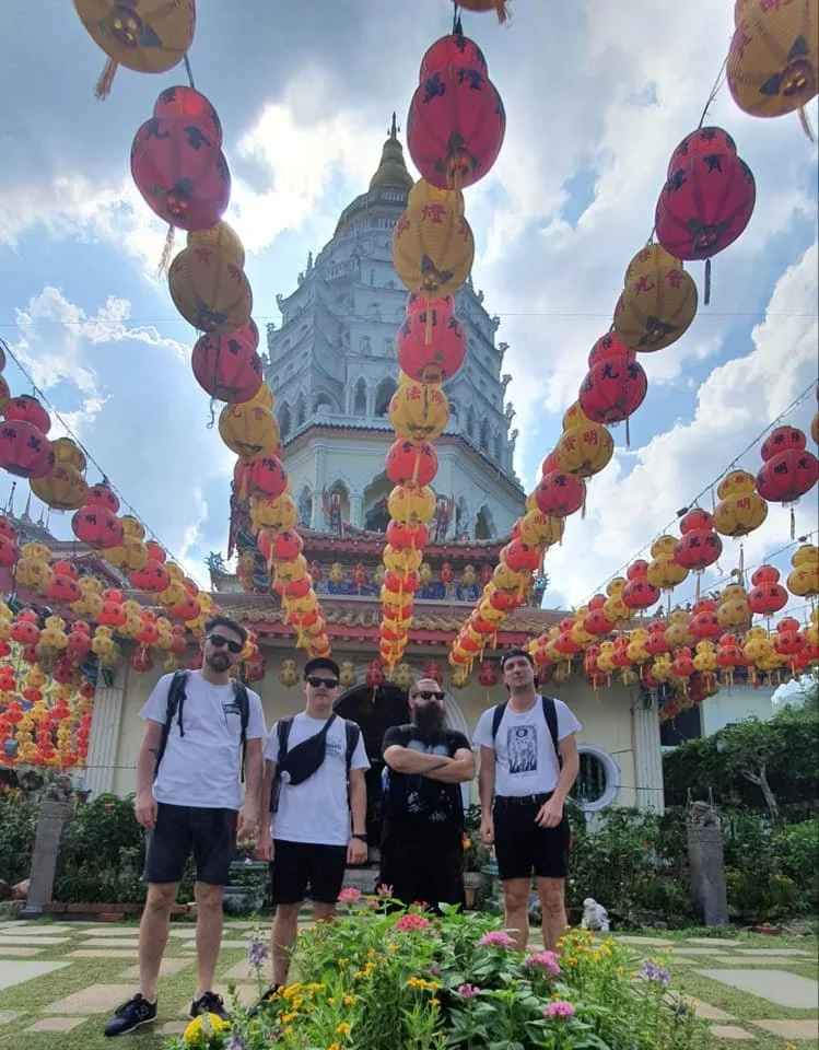 Four men with backpacks standing in front of a decorated Asian temple with hanging red and yellow lanterns and a garden in front.